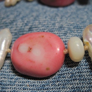 May include: A close-up of a pink and white beaded necklace. The beads are made of coral and pearl. The necklace is lying on a blue denim fabric.
