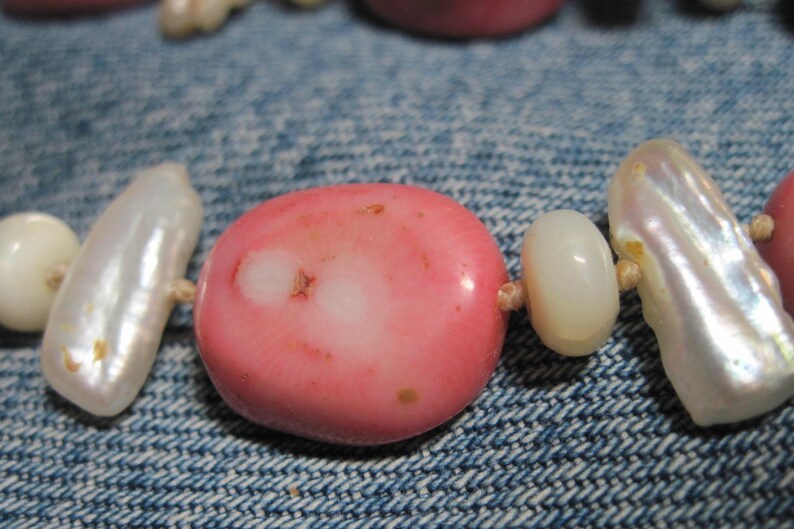 May include: A close-up of a pink and white beaded necklace. The beads are made of coral and pearl. The necklace is lying on a blue denim fabric.