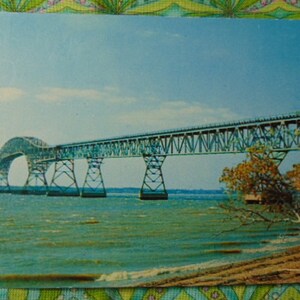 May include: A vintage postcard featuring a photo of a large, steel bridge over a body of water. The bridge is light blue and has a curved section in the middle. The water is a light blue-green color. The sky is a light blue color with some clouds. There are trees on the right side of the image.