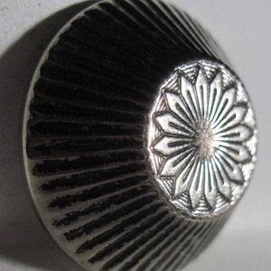May include: A silver-toned metal button with a decorative, radial pattern and a raised, stylized flower design in the center.