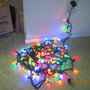 May include: A tangled string of Christmas lights with multi-colored bulbs, including red, green, blue, yellow, and orange. The lights are plugged into an outlet and are lying on a light brown floor. The date 03/08/2017 is written on the floor.