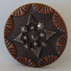 May include: A brown and silver button with a starburst design. The button has a textured surface and a raised design.