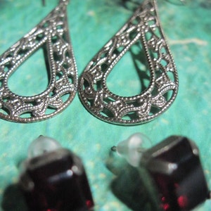 May include: A pair of silver filigree earrings with a teardrop shape and a dark red gemstone stud earring.