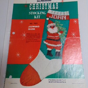 May include: A Bucilla Christmas stocking kit package featuring a jumbo size stocking with a Santa Claus graphic holding a tree. The design includes the name JOAN, red and green color blocks, white accents, and star motifs. Text includes NO. 7621.