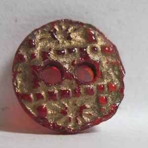 May include: A red glass button with a gold pattern and two holes. The button has a checkered pattern and a raised design.