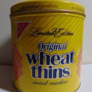 May include: A yellow, cylindrical tin of "Original Wheat Thins" snack crackers. The tin features the Nabisco logo and the words "Limited Edition." The text "whole wheat goodness" is printed on the lid. The net weight is 13 oz.
