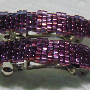 May include: Two purple iridescent hair clips with a silver metal clasp. The clips are made of small square beads.