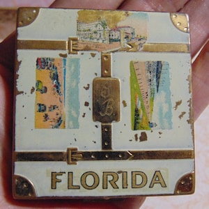 May include: A white metal compact with gold trim and a vintage postcard design. The compact has a hinged lid and a mirror inside. The postcard design features images of Florida and the text "Florida".