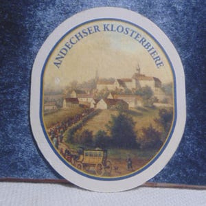 May include: A round cardboard beer coaster featuring a printed illustration of a village with a monastery on a hill, a horse-drawn carriage, and a procession of people. The text ANDECHSER KLOSTERBIERE is printed in blue along the top edge.