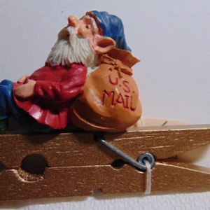 May include: A whimsical clothespin featuring a seated gnome figurine. The gnome wears a blue hat, red jacket, and blue pants, with a white beard. A tan satchel with "U.S. MAIL" is next to him. The clothespin is gold-colored.