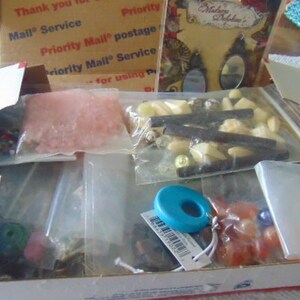 May include: A cardboard box filled with various craft supplies, including beads, sequins, and small plastic pieces. The box is open and the supplies are visible in clear plastic bags.