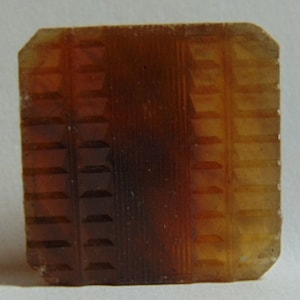 May include: A square, translucent, amber-colored glass tile with a textured, geometric pattern.
