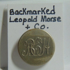 May include: A silver button with the initials "LM" in a cursive script. The button is labeled "Backmarked Leopold Morse + Co."