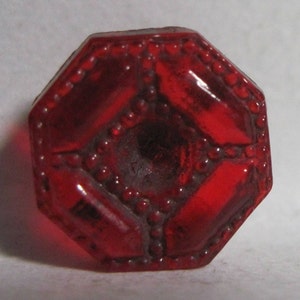 May include: A red glass octagonal button with a raised, textured design. The button has a small hole in the center.