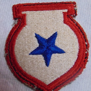 May include: A white patch with a red border and a blue star in the center. The patch is shaped like a shield.