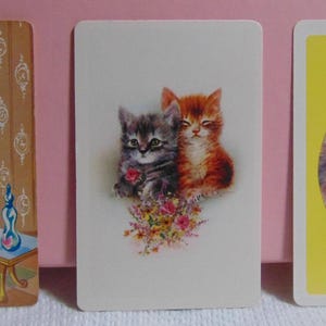 May include: Three vintage cards featuring illustrations of cats. The first card shows a Siamese cat sitting on a red chair with gold accents. The second card shows two cats, one grey and one orange, sitting together with a bouquet of flowers. The third card shows a brown tabby cat sitting on a yellow background.