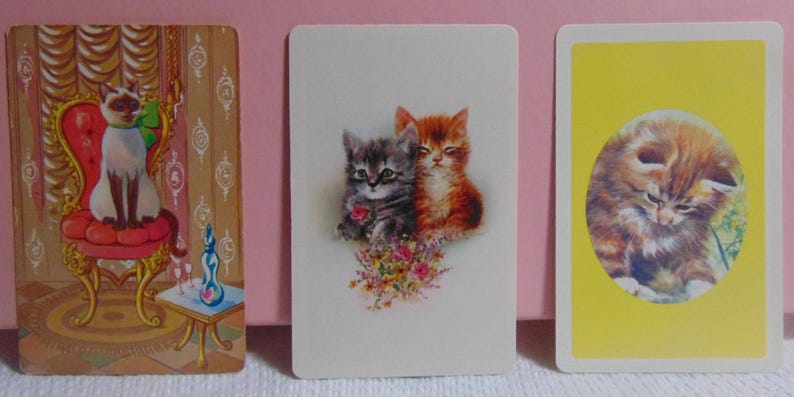 May include: Three vintage cards featuring illustrations of cats. The first card shows a Siamese cat sitting on a red chair with gold accents. The second card shows two cats, one grey and one orange, sitting together with a bouquet of flowers. The third card shows a brown tabby cat sitting on a yellow background.