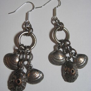 May include: A pair of silver-toned dangle earrings featuring multiple metal seashell charms suspended from a circular ring. Each earring includes three distinct shell-shaped pendants with textured surfaces and a standard hook-style ear wire closure.