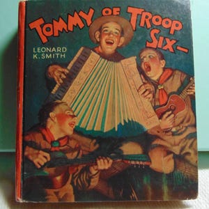 May include: A vintage book cover featuring three boys in scout uniforms playing musical instruments. The book title is "Tommy of Troop Six" by Leonard K. Smith. The cover art is illustrated in a colorful style with a red, white, and blue color scheme.
