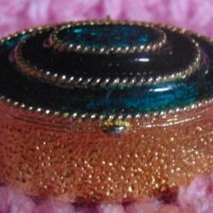 May include: A small, oval-shaped trinket box with a gold-colored metal exterior and a teal-colored enamel lid. The lid has a raised, circular design with a gold trim.