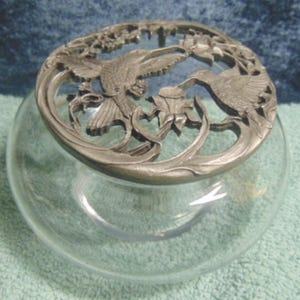 May include: A clear glass bowl topped with a circular, silver-toned metal lid featuring an intricate openwork design of two hummingbirds hovering near flowers. The metal lid rests securely on the rim of the rounded, transparent glass vessel.