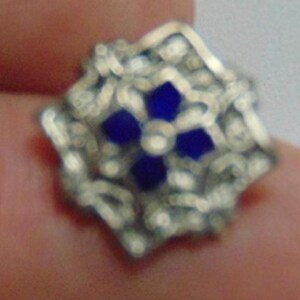 May include: A silver-toned, square-shaped jewelry piece with an ornate design. The center features a four-petal flower design in a deep blue color. The piece is held between fingers.