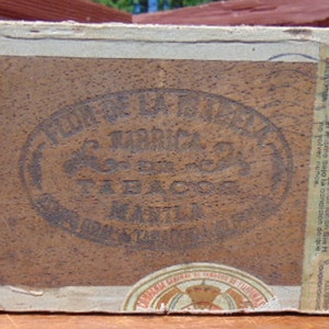 May include: A vintage wooden box with a brown leather label that reads "Flor de la Isla" and "Tabacos Manila". The box has a warning label in Spanish and English.