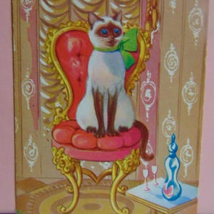 May include: A Siamese cat with blue eyes sits on a red velvet chair with gold trim. The cat is wearing a green bow. The chair is in a room with gold wallpaper and a small table with a bottle and glasses.