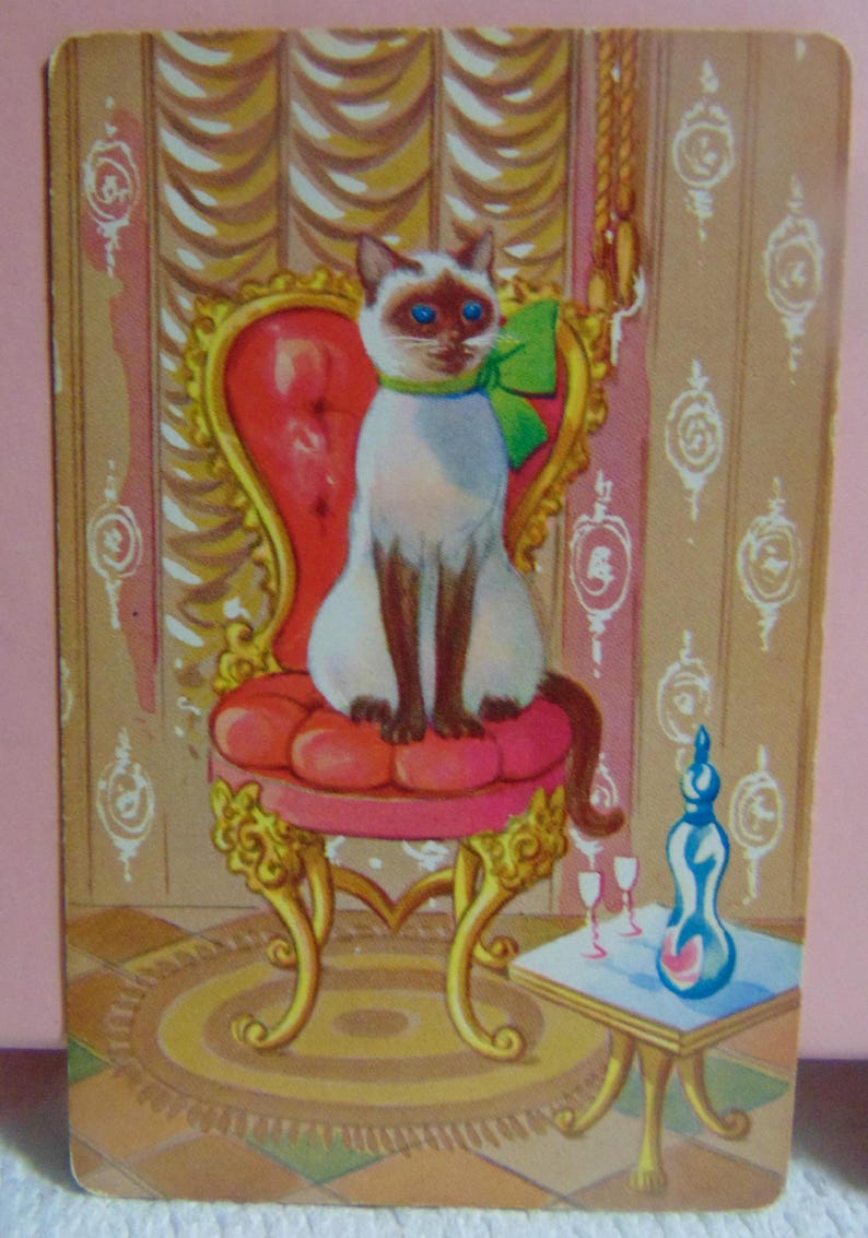 May include: A Siamese cat with blue eyes sits on a red velvet chair with gold trim. The cat is wearing a green bow. The chair is in a room with gold wallpaper and a small table with a bottle and glasses.
