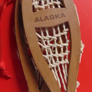 May include: A pair of wooden snowshoes with a white woven net, the word "ALASKA" is carved into the wood.