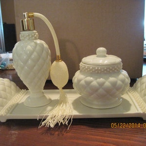 May include: A white porcelain perfume bottle set with a matching jar and tray. The bottle has a gold top and a white tassel. The jar has a white lid with a pearl trim. The tray has a pearl trim and a quilted design.