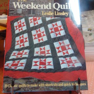 May include: A book titled "The Weekend Quilt" by Leslie Linsley. The cover features a quilt design with red and white star patterns on a dark gray background, set against a red border. The book is displayed on a metal stand.