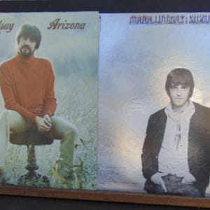 May include: Two vinyl record album covers featuring Mark Lindsay. The left cover, titled Arizona, shows a person in an orange turtleneck and light pants sitting in a field. The right cover, Mark Lindsay and Silver Bird, shows a person in a dark jacket.