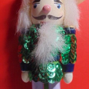 May include: A wooden nutcracker ornament with a blue and white hat, green and gold sequined jacket, white beard, and black shoes. The ornament is standing on a green base.