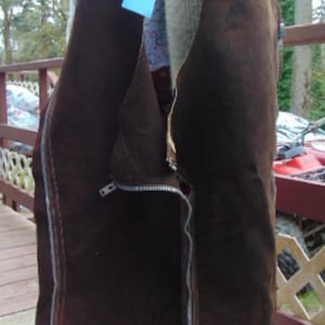 May include: Brown suede chaps with a silver zipper and a belt. The chaps are held up by a person. A blue tag hangs from the belt. The chaps appear to be vintage.