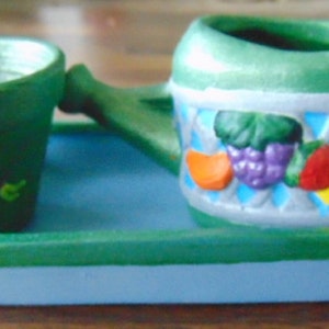 May include: A green ceramic tray with a blue base holds a green ceramic watering can with a fruit design and a smaller green ceramic pot.