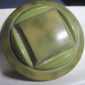 May include: A green and brown plastic button with a square design in the center. The button has a raised edge and a smooth surface.