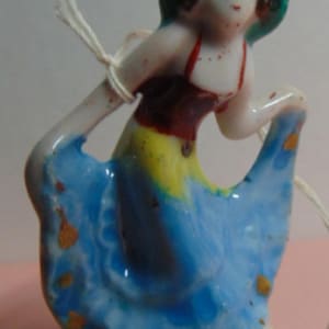 May include: A small, vintage ceramic figurine of a woman in a blue dress with gold accents. The figurine has a green hat and a red top. The dress has yellow and red details. A white string is tied around the figurine.