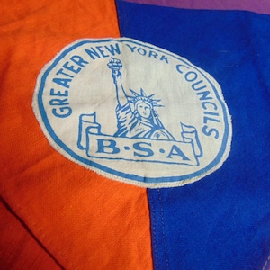 May include: A patch with the text "GREATER NEW YORK COUNCILS B.S.A." and an illustration of the Statue of Liberty. The patch is white with blue text and a blue border. The background is orange, blue, and purple fabric.