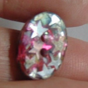 May include: Oval-shaped button with a pink, iridescent, and white finish. The button is covered in small, white stars.