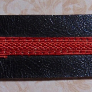 May include: A black leather strap with a red woven ribbon sewn across the center. The strap has two holes on either side.