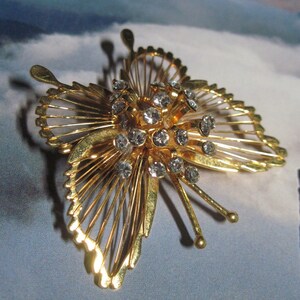 May include: A gold-tone butterfly brooch with a cluster of clear rhinestones in the center. The wings are made of delicate wire and have a fan-like design.