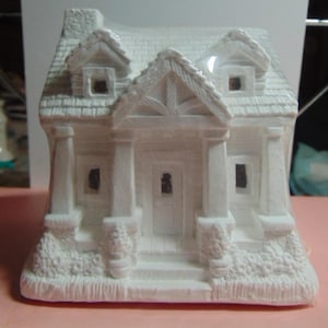 May include: A white ceramic miniature house with a detailed facade. The house features a front porch with columns, a front door, windows, and a textured roof. The house has a chimney and decorative floral accents at the base.