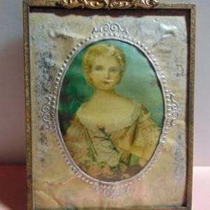 May include: Antique portrait in a rectangular frame with an ornate gold-colored top. The oval portrait features a woman in a light-colored dress. The frame has a silver-colored oval border around the portrait and a gold-colored frame.