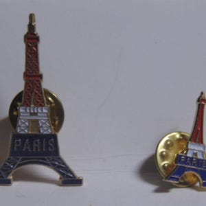 May include: Two metal lapel pins shaped like the Eiffel Tower, each featuring red, white, and blue enamel detailing. The base of each pin displays the word PARIS in raised gold lettering against a dark blue background, with gold metal pin backs.