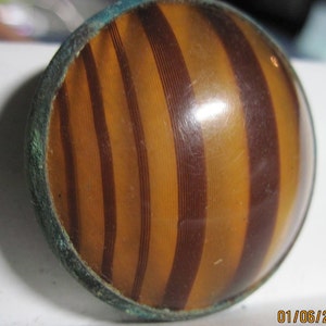 May include: A round, striped, brown and orange glass button with a metal rim.