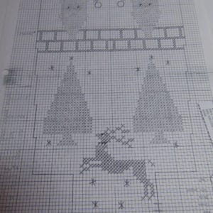 May include: A cross-stitch pattern printed on white grid paper featuring two figures wearing Santa hats positioned above a brick wall. Below the wall are two triangular evergreen trees and a leaping reindeer, all rendered in dark blue ink.