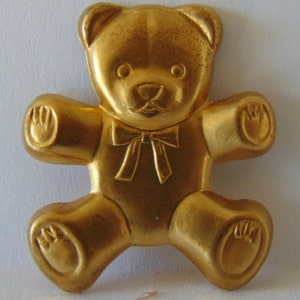 May include: A gold-colored teddy bear figurine with a bow tie. The bear is sitting with its arms outstretched and its feet are visible.