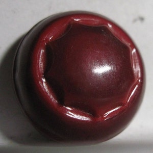 May include: A dark red button with a raised, octagonal design. The button is smooth and shiny.