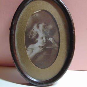 May include: Oval-shaped antique picture frame with a dark brown wooden frame. The frame holds a sepia-toned photograph of a child. The photograph is surrounded by a gold-colored matting. The frame is approximately 6 inches tall.